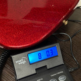 A close-up of a red glitter-finish guitar body resting on a digital smart weigh scale displaying 8.03 kg, set on a dark wood surface.