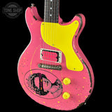 A pink electric guitar with a bright yellow pickguard, distressed finish, and a circular black-and-white portrait graphic on the lower bout. The neck is black with white dot inlays.