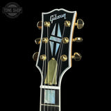 Close-up of a Gibson electric guitar headstock with gold tuning pegs, a black faceplate, and mother-of-pearl inlays on a black background.