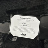 A close-up of a Gibson Custom Certificate of Authenticity card inside a black case, placed on a black furry surface. The card is white with black text and the Gibson Custom logo at the bottom.