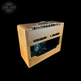 A vintage-style guitar amplifier with a tan textured fabric cover, black control knobs, and a metal handle, shown against a solid black background with a Tone Shop watermark in the top-left corner.