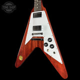 A redwood V-shaped electric guitar with a white pickguard, black pickups, and black control knobs, shown against a solid black background with a Tone Shop watermark in the top left corner.