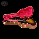 An acoustic guitar with a sunburst finish displayed inside an open brown hard case with a bright pink velvet lining, set against a solid black background.