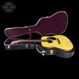 An open acoustic guitar case with a natural wood-finish acoustic guitar resting inside, photographed against a solid black background. A Tone Shop watermark is visible in the top-left corner.