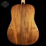 Close-up of the back of an acoustic guitar showing a rich, warm brown wood grain finish, set against a solid black background with a Tone Shop watermark in the top left corner.