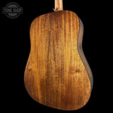 Close-up of the back of an acoustic guitar showing a rich, warm brown wood grain finish, set against a solid black background with a Tone Shop watermark in the top left corner.