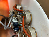 Close-up of an electronic guitar control module showing silver potentiometers, orange and blue wires, and a white plastic housing.