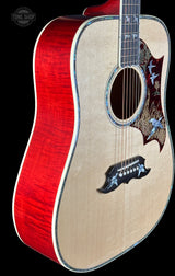 Close-up of an acoustic guitar body showing a bright red cherry wood back, natural wood top, decorative bird inlay on the soundboard, and a black pickguard, photographed against a black background.