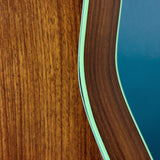 Close-up detail of a guitar body showing a warm brown wood grain finish, a curved edge with a pale green binding, and part of a blue soundhole surround.