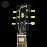Close-up of a Gibson acoustic guitar headstock against a black background, showing the Gibson logo, tuning pegs, strings, and part of the neck.