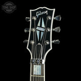 Close-up of a Gibson electric guitar headstock against a black background, showing the Gibson logo, six tuning pegs, strings, and part of the fretboard.