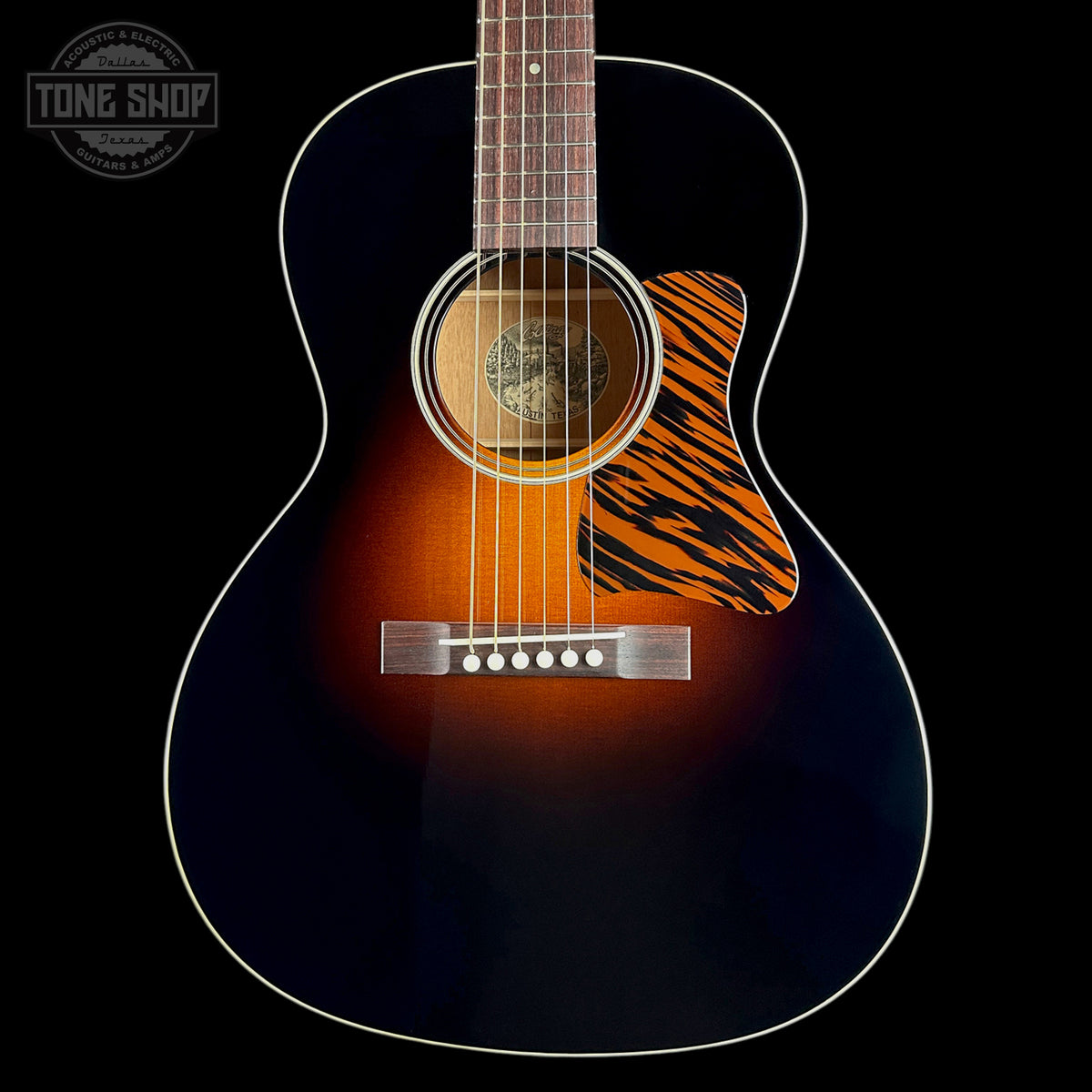 A close-up studio shot of an acoustic guitar body against a black background, showing a dark blue to brown sunburst finish, white binding, a tiger-stripe style pickguard, and a silver bridge. A Tone Shop watermark is visible in the top left corner.