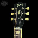 Close-up of a Gibson acoustic guitar headstock with six light yellow tuning pegs, gold-toned tuners, and a black faceplate on a black background.