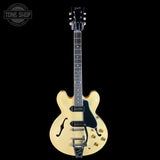 An electric guitar with a cream-colored body, black pickguard, and gold hardware, shown against a solid black background. The Gibson logo is visible on the headstock, and a Tone Shop watermark is in the top-left corner.
