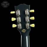 Close-up of the headstock of a Gibson electric guitar against a black background, showing six silver tuning machines with pale yellow tuning keys and a gold Gibson logo on the black neck.