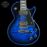 A close-up of a blue electric guitar body with a glossy flamed maple finish, black pickguard, gold hardware, and silver control knobs, shown against a black background with a Tone Shop watermark in the top left.