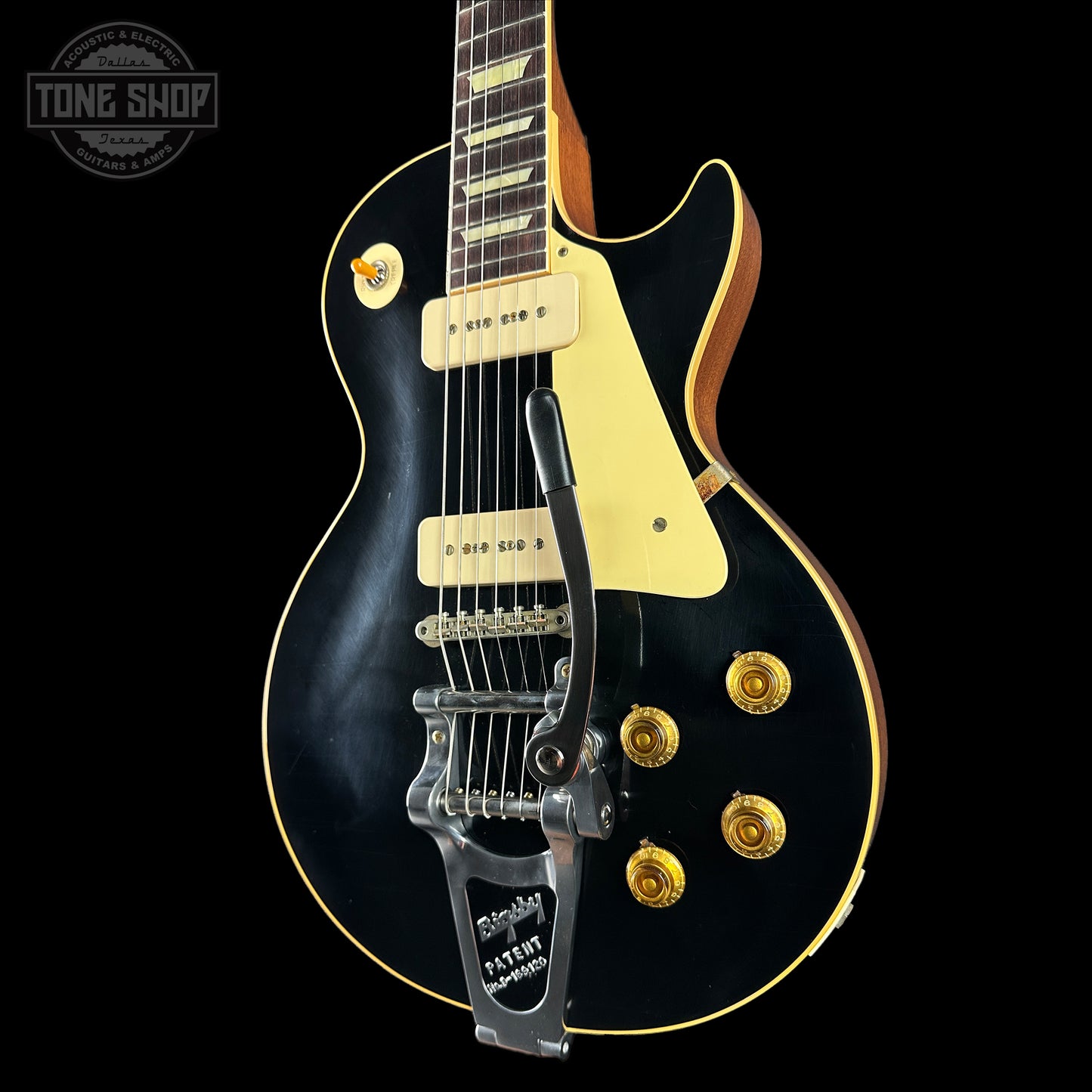 Black electric guitar with gold hardware on a black background, featuring 'Tone Shop' logo.