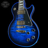 A blue electric guitar with a glossy blue flame finish, black pickguard, and gold hardware, shown against a black background with a Tone Shop watermark in the top left corner.