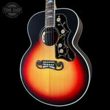 A close-up studio shot of an acoustic guitar body against a black background, showing a sunburst finish that transitions from bright yellow-orange in the center to deep red and black around the edges. The guitar features a black pickguard with gold and white floral inlays, a decorative black bridge, and a dark fretboard.