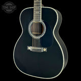 A close-up studio shot of an acoustic guitar body against a black background, featuring a glossy black finish, white binding, and a silver bridge. A Tone Shop watermark is visible in the top-left corner.
