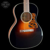 A close-up studio shot of an acoustic guitar body against a black background, showing a dark brown to black sunburst finish, white binding, a tiger-stripe style pickguard, and part of the neck with a light wood fretboard. A Tone Shop watermark is visible in the upper left corner.