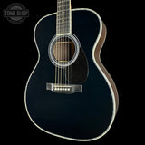 A close-up studio shot of an acoustic guitar body against a black background, showing a glossy black finish, white binding, a dark pickguard, and silver-toned hardware. A Tone Shop logo watermark is visible in the top-left corner.