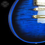 Close-up of a blue electric guitar body with a flamed maple wood finish, gold hardware, and a black background. A Tone Shop watermark is visible in the top left corner.