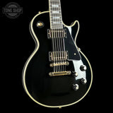 Black electric guitar with gold hardware on a black background, featuring 'Tone Shop' logo.