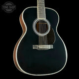 A black acoustic guitar with a glossy black finish, white rosette detailing, and a dark brown wooden body edge, shown against a solid black background. A Tone Shop watermark is visible in the top-left corner.