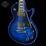 A blue electric guitar with a glossy blue flame finish, black pickguard, and gold hardware, shown against a black background with a Tone Shop watermark in the top left corner.