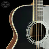 Close-up of an acoustic guitar body showing the black glossy finish, natural wood edges, and decorative rosette around the sound hole, with part of the fretboard and strings visible against a black background.