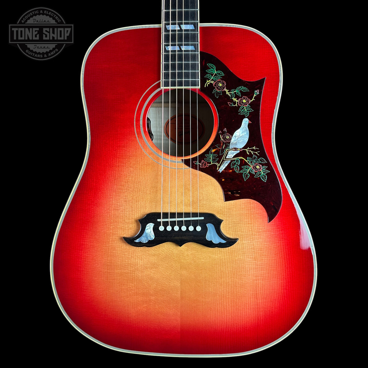 A front-facing acoustic guitar with a red-to-tan sunburst finish, featuring a dark pickguard with a white bird and floral inlay, set against a solid black background.