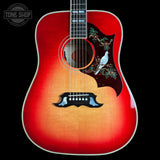 A front-facing acoustic guitar with a red-to-tan sunburst finish, featuring a dark pickguard with a white bird and floral inlay, set against a solid black background.