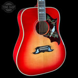 A red acoustic guitar with a natural wood sunburst finish, featuring a decorative black pickguard with a white bird and green floral inlay, shown against a black background.