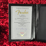 A Fender Certificate of Authenticity card inside a black leather-bound document holder, placed on a red textured fabric background.