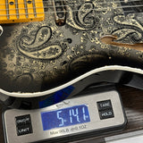 A close-up of an electric guitar body with a black and beige paisley pattern, resting on a digital scale displaying 5.141 lbs.