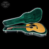 An acoustic guitar with a natural wood finish and black pickguard, displayed inside an open green-lined hard case on a black background. A Tone Shop watermark is visible in the top-left corner.