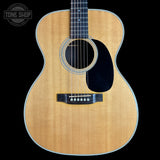 An acoustic guitar with a natural wood finish and black pickguard, shown against a solid black background with a Tone Shop watermark in the top left corner.
