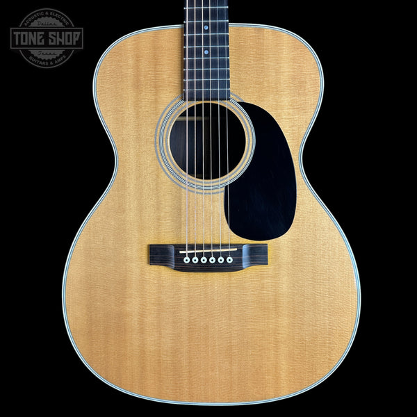 An acoustic guitar with a natural wood finish and black pickguard, shown against a solid black background with a Tone Shop watermark in the top left corner.