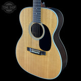 An acoustic guitar with a natural wood top, dark sides, and a black pickguard, shown against a solid black background with a Tone Shop watermark in the top-left corner.