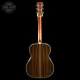 The back of an acoustic guitar with a dark brown wood grain finish, shown against a solid black background. A Tone Shop watermark is visible in the top-left corner.
