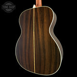 Close-up of the back of an acoustic guitar with a dark brown wood grain finish and a light cream binding, shown against a black background with a Tone Shop watermark in the top left.