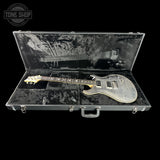 An electric guitar with a silver flamed maple body and gold-toned headstock, displayed inside an open black hard case with a black plush interior. A Tone Shop watermark is visible in the top-left corner.
