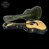 An acoustic guitar with a natural wood top and black headstock, resting inside an open black hard case with green velvet lining, shown against a solid black background. A Tone Shop watermark is visible in the top-left corner.