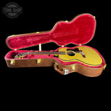 An acoustic guitar with a natural wood top and dark neck, displayed inside an open brown hard case lined with bright pink velvet. The case is photographed against a solid black background, with a Tone Shop watermark in the upper left corner.