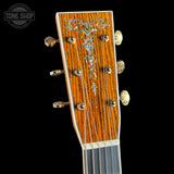 Close-up of an acoustic guitar headstock with a warm orange-brown wood grain finish, ornate silver and blue inlay, six gold-toned tuning pegs, and six strings, shown against a black background with a Tone Shop watermark.