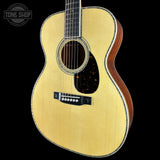 A close-up of a wooden acoustic guitar body with a natural light wood finish, black pickguard, and dark fretboard, shown against a solid black background with a Tone Shop watermark in the top-left corner.
