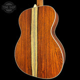 Close-up of the back of an acoustic guitar showing a rich reddish-brown wood body with a central light wood inlay, set against a solid black background. A Tone Shop watermark is visible in the top-left corner.