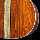 Close-up detail of an acoustic guitar body showing rich warm brown wood grain, a light natural wood inlay stripe, and a silver metal edge against a black background.