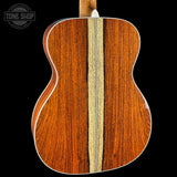 Close-up of the back of an acoustic guitar showing a rich reddish-brown wood grain finish with a central light wood strip, photographed against a solid black background. A Tone Shop watermark is visible in the top-left corner.